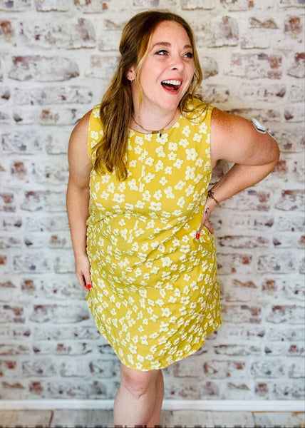 Woman in a yellow floral dress standing against a brick wall.