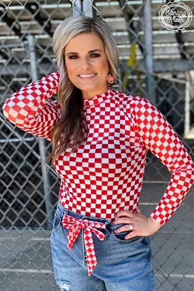 A woman wearing a red and white checkered mesh top with three-quarter length sleeves, paired with blue denim shorts, posing with one hand on her head.