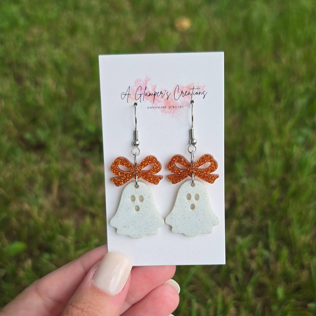 Earrings with ghost designs and orange bows on a card held against a grassy background.