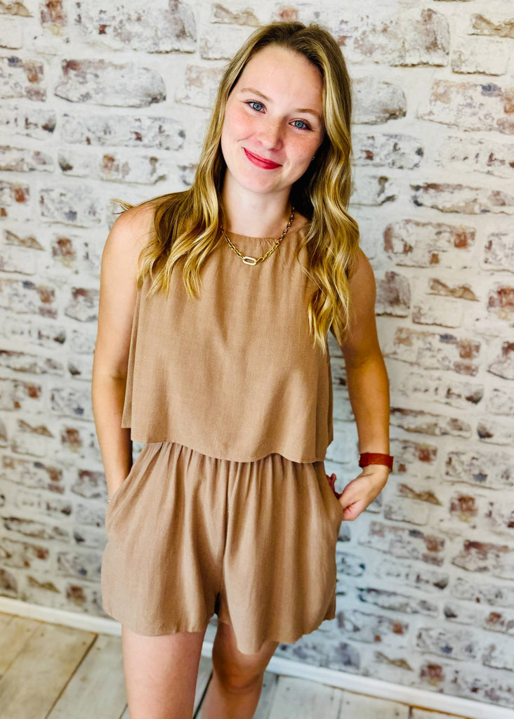 Woman wearing a beige romper against a brick wall.