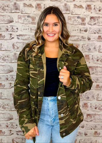 A woman wearing a green camouflage jacket over a black top and light blue jeans, standing in front of a white brick wall.